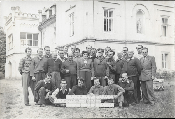 Na fotografii je 25 uitel a jedna pan nebo slena uitelka na trvnku a cest ped koln budovou. Pan nebo slena uitelka 
    je uprosted v pedn stojc ad. Vpravo za nimi vykukuje korek se sedcm dttem. Ped nimi je poloeno logaritmick pravtko 
    o velkch rozmrech. slice na pravtku nejsou pi tchto rozmrech fotografie iteln.